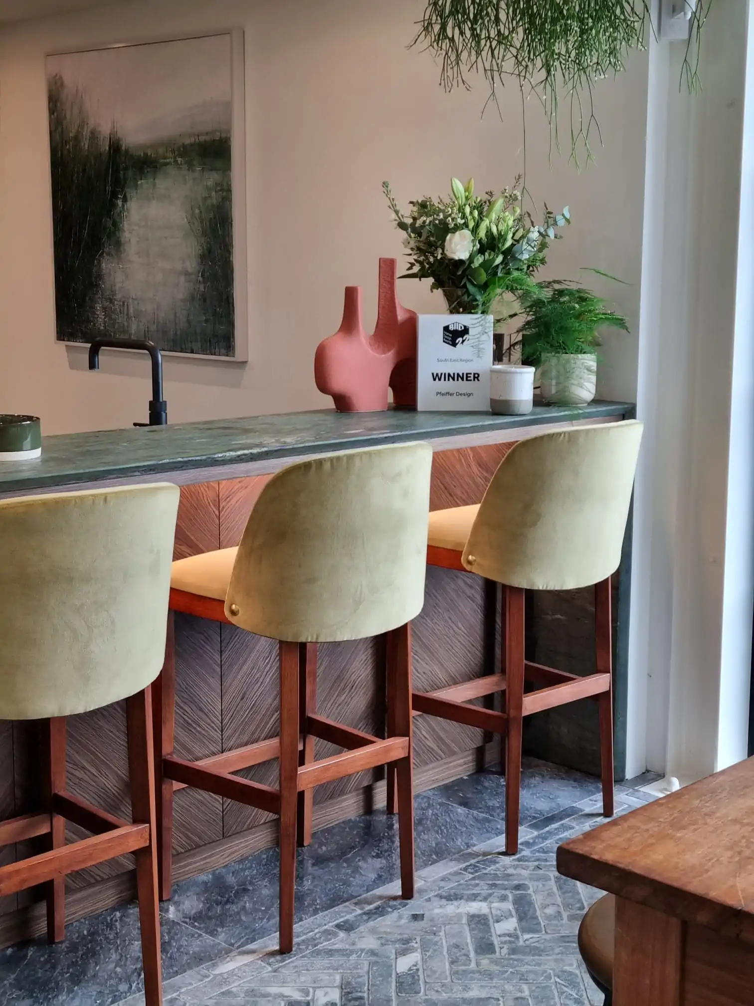 Three yellow-green bar stools are positioned at a wooden counter with a textured backdrop. A plant arrangement and a decorative vase are atop the counter, amidst natural lighting and a modern setting. A sign reads: "WINNER Pfeffer Design."
