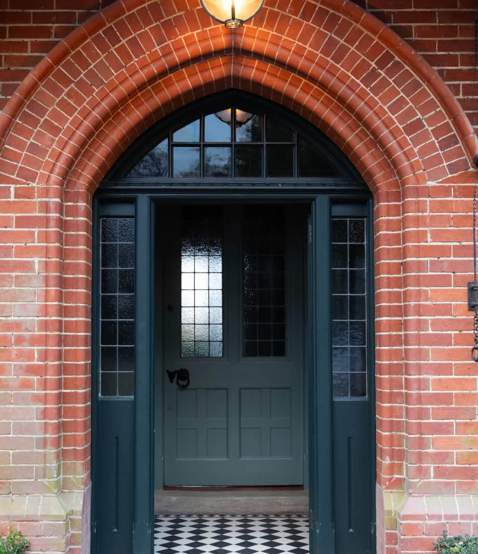 A green double door stands open, framed by a curved red brick arch. Glass panels alongside the door reflect light, while a patterned black and white tiled floor is visible inside.