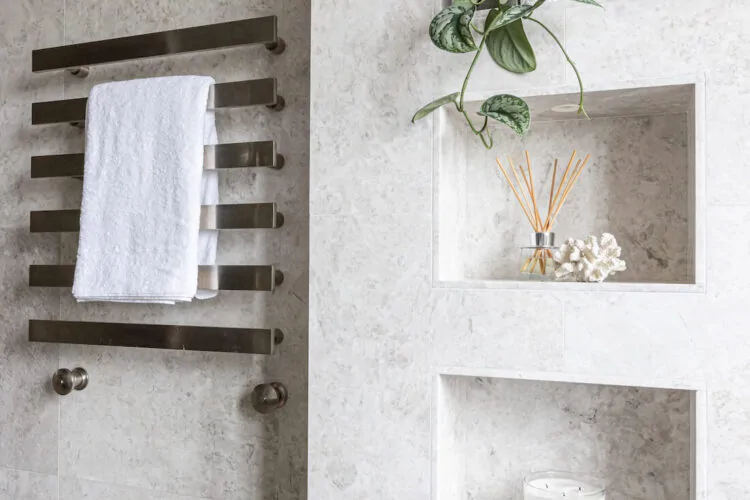 A sleek bathroom wall features a towel rack holding a white towel and showcases stylish niches with a plant, decorative items, and a candle, all set against a light stone backdrop.