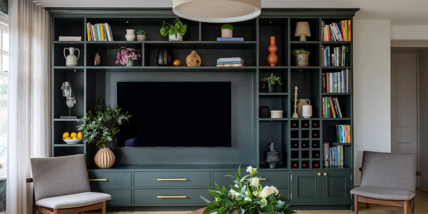 A green bookshelf with decorative items and a television is mounted centrally. Two armchairs face a wooden coffee table adorned with a floral arrangement and magazines in a bright room.