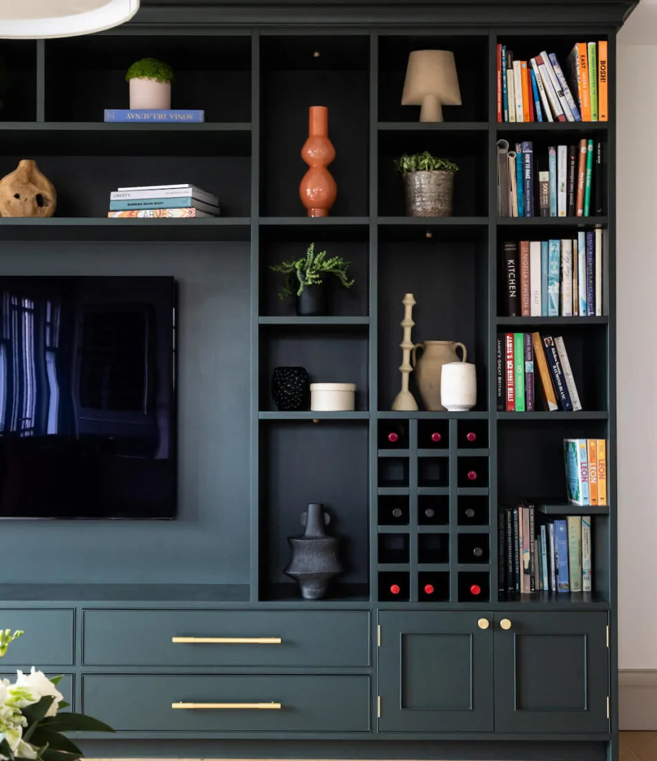 A dark green shelving unit displays various decorative items, including books, vases, and plants, alongside a television, against a light wall and wooden flooring.
