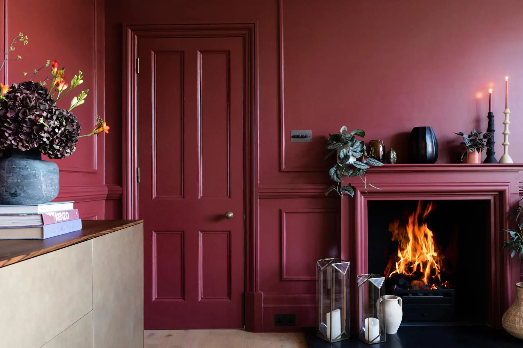 A dark-red wall features a door and a fireplace with flames. A wooden sideboard holds books, a flower vase, and decorative items. Candles glow atop the mantle, surrounded by plants.