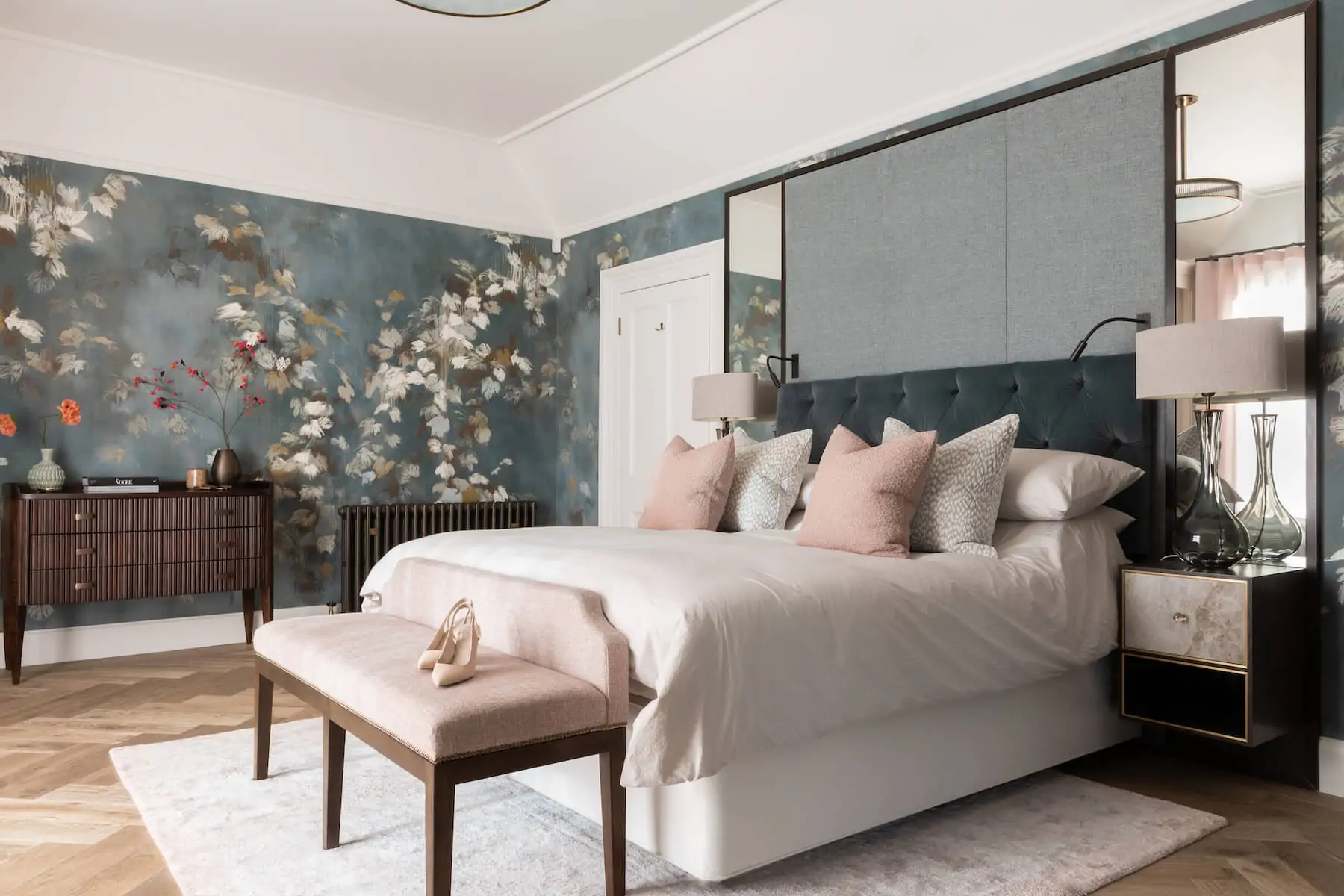 A stylish bedroom features a large bed with decorative pillows, a bench with a single shoe, and a floral wallpaper backdrop. Soft lighting illuminates a modern dresser and nightstand.