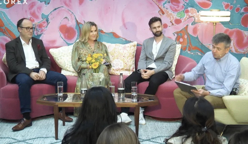 Four individuals are seated on a pink couch, engaged in discussion. One holds a notebook and flowers, while a table with glasses of water sits in front of them. A colorful abstract background sets the scene.