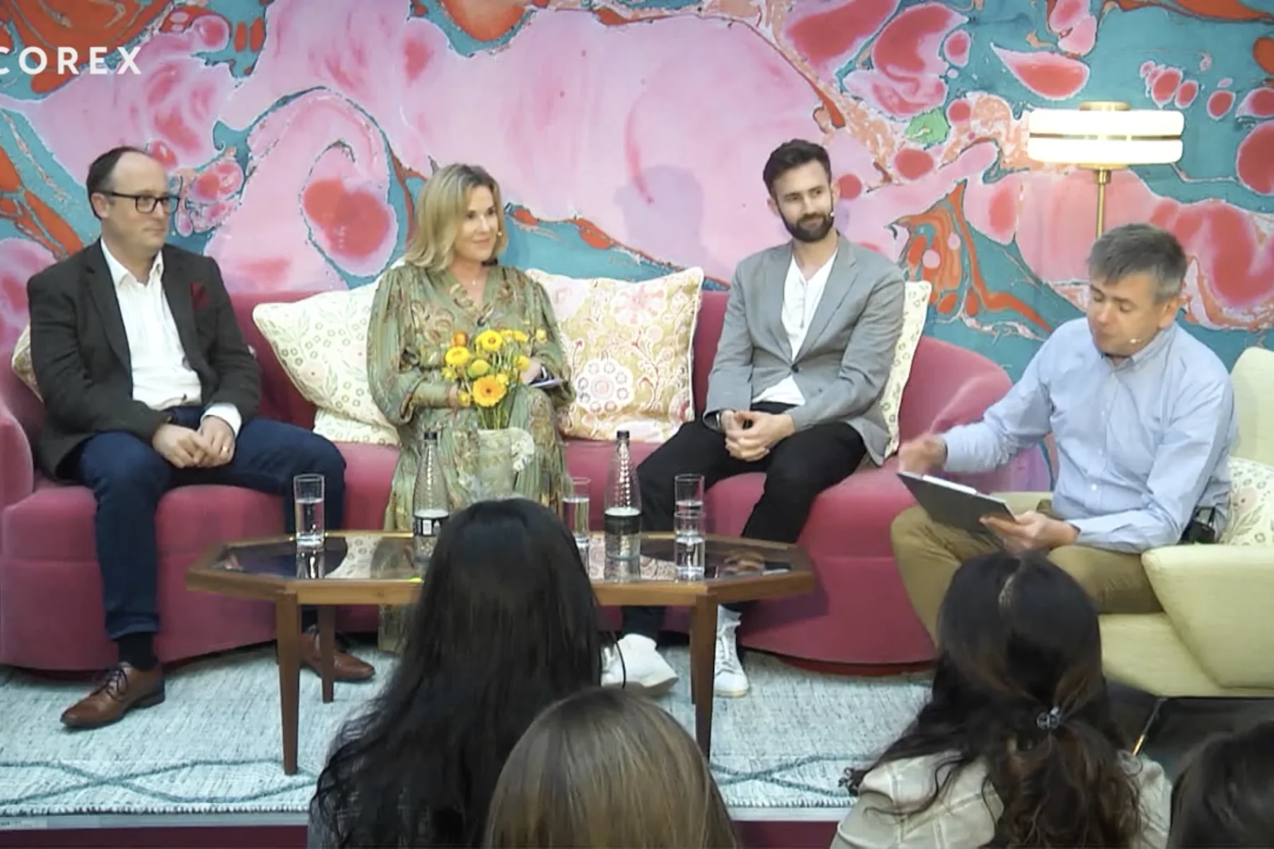 Four individuals are seated on a pink couch, engaged in discussion. One holds a notebook and flowers, while a table with glasses of water sits in front of them. A colorful abstract background sets the scene.