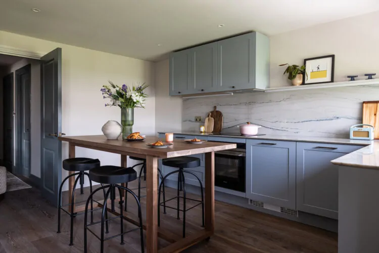 A modern kitchen features a wooden table with black stools, plates of food, and a vase of flowers. The light gray cabinetry complements the marble backsplash, creating a bright and inviting atmosphere.