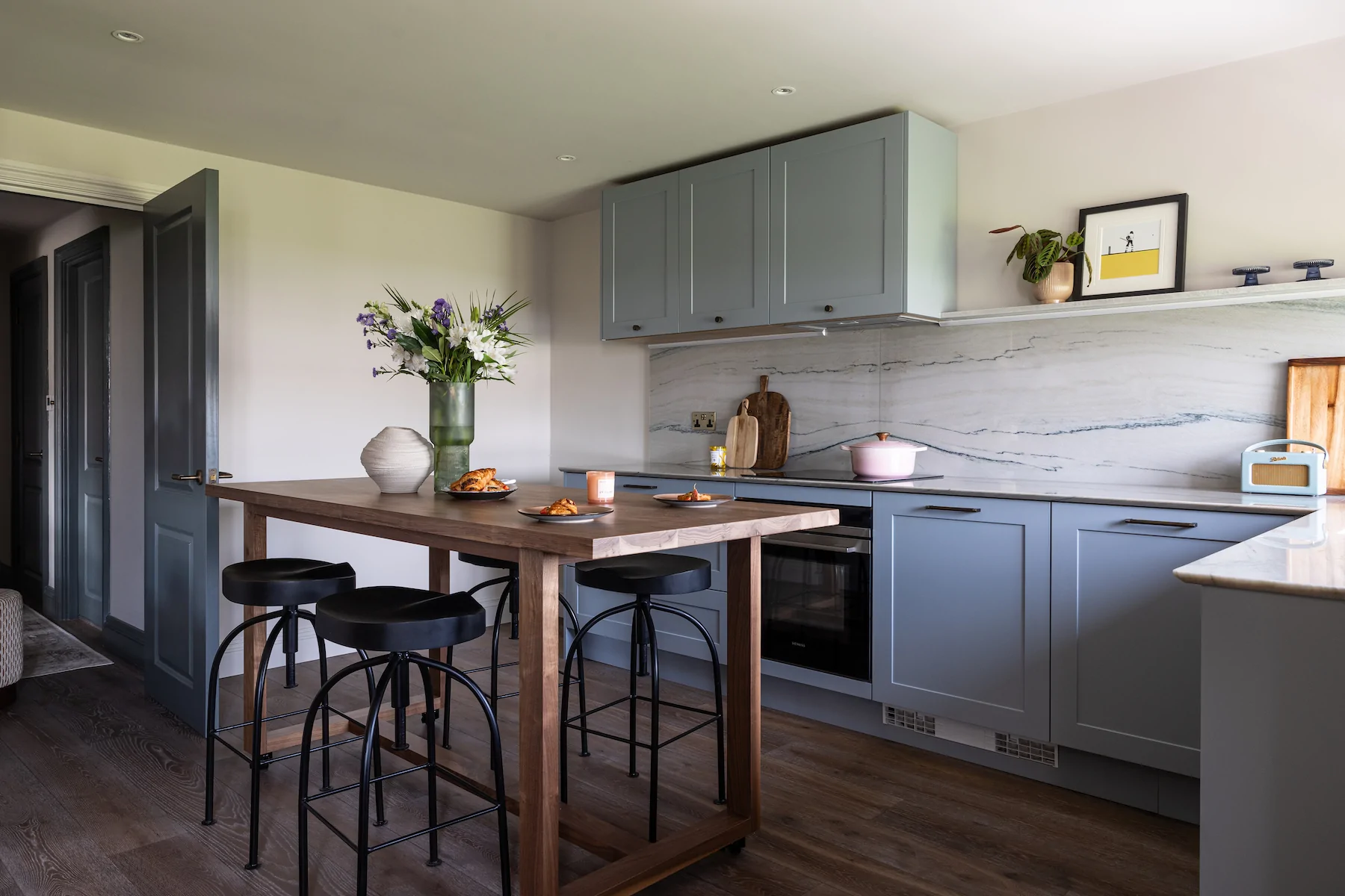 A modern kitchen features a wooden table with black stools, plates of food, and a vase of flowers. The light gray cabinetry complements the marble backsplash, creating a bright and inviting atmosphere.