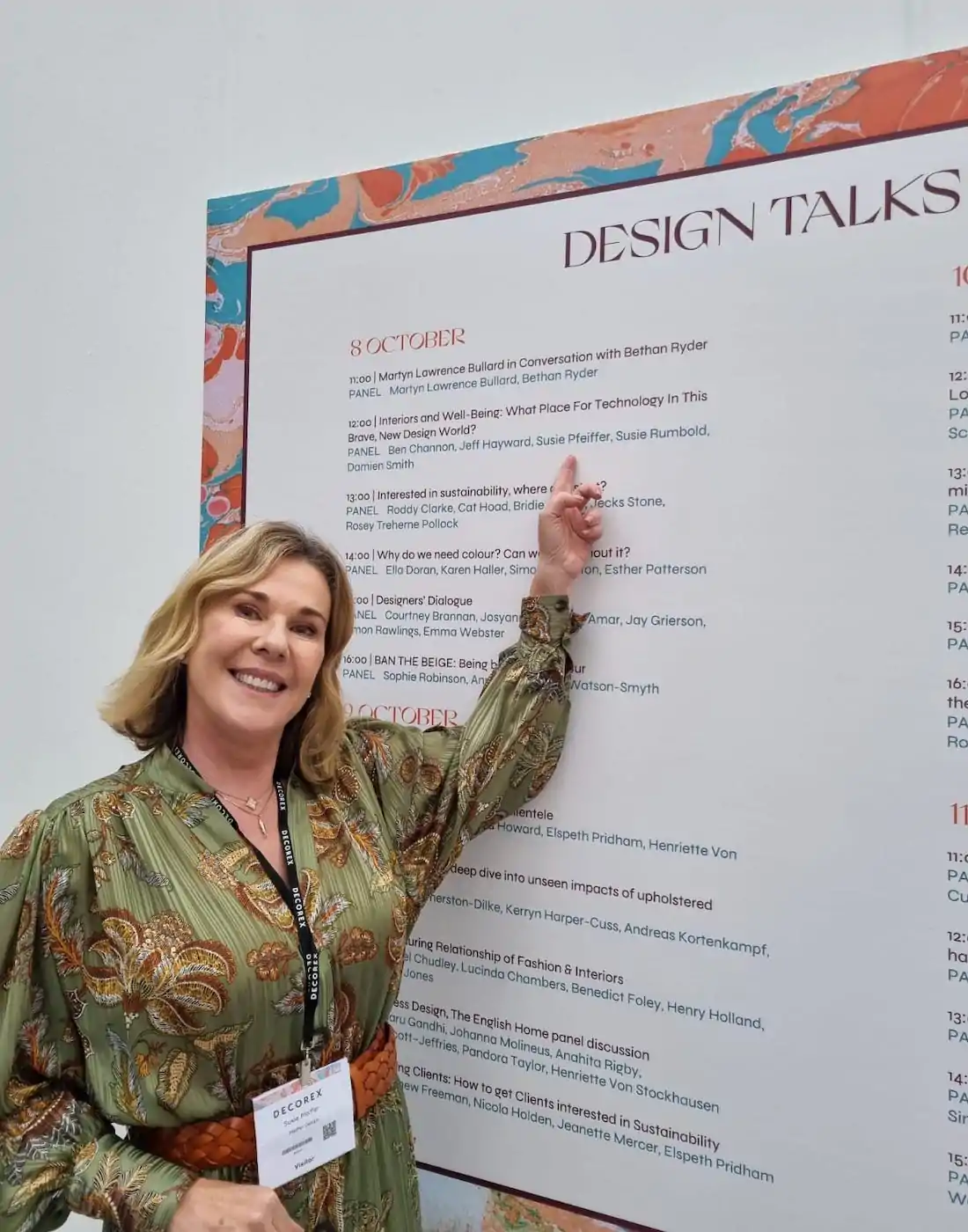 A woman in a patterned green dress points at a schedule titled "DESIGN TALKS," detailing events for October 8th, against a decorative wall backdrop. She smiles, wearing a name tag.