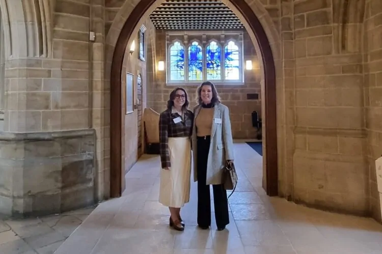 Two women stand smiling together in a stone archway. Behind them is a bright stained glass window, and the interior features a mix of rustic and modern elements.