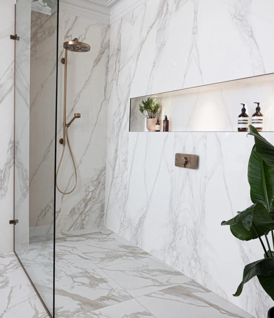 A marble-tiled bathroom features a glass shower with a bronze showerhead. In a recessed shelf, plants and decorative bottles sit, while a potted plant adds greenery to the space.
