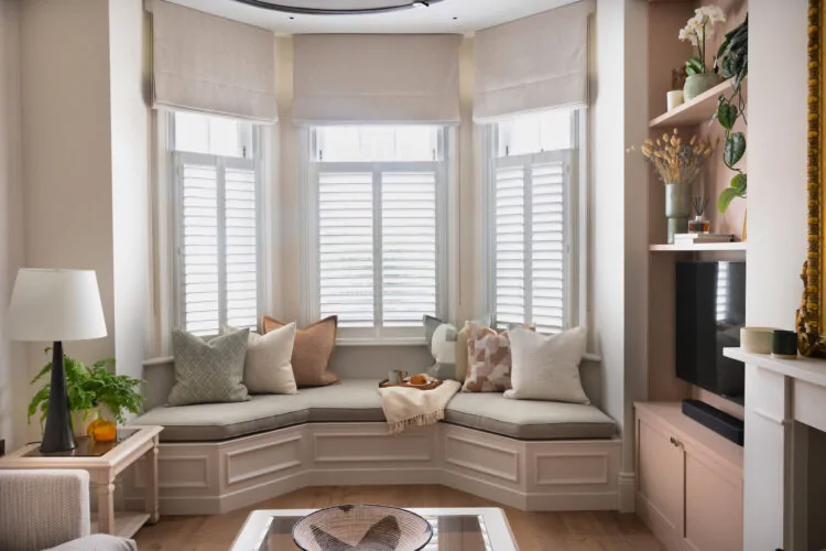 A cosy window nook features plush pillows and a blanket, while a lamp and plant adorn a nearby table. Light filters through shuttered windows, enhancing the warm ambiance.