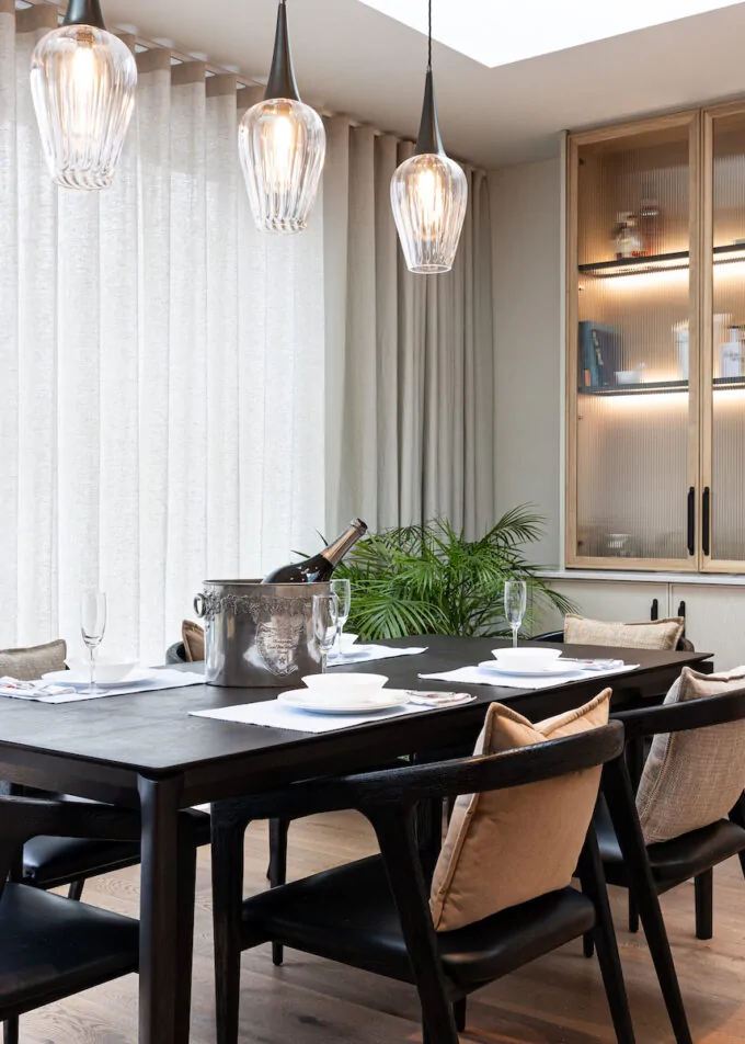 A black dining table is set with plates and glasses, holding an ice bucket with a bottle. Large windows with sheer curtains let in light, surrounded by modern decor and plants.