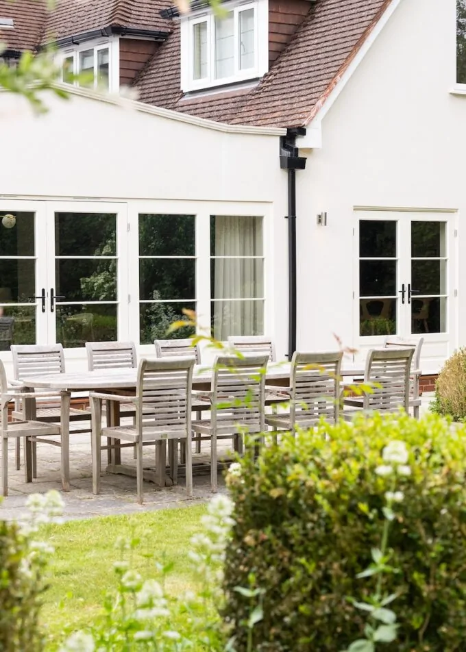 A wooden dining table with chairs is set outside a light-colored house. The scene is surrounded by manicured shrubs and colorful flowers, suggesting a serene garden setting.