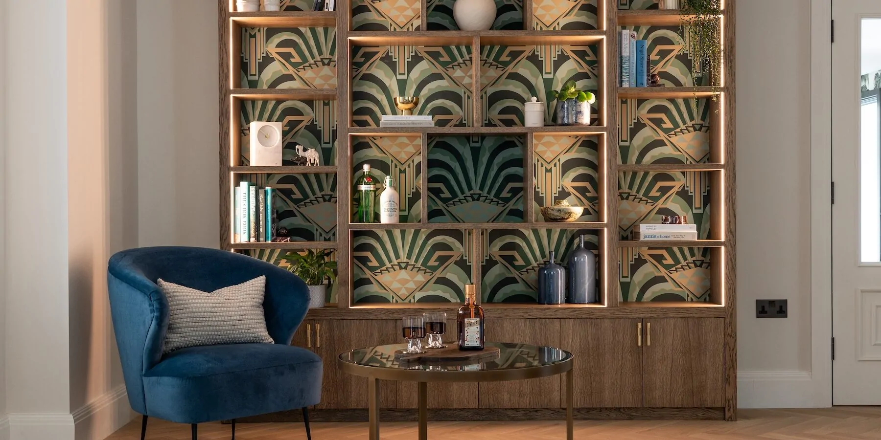Art Deco Inspired Bespoke Bar and Library made by our bespoke joiners, designed by Pfeiffer Design. Art deco wallpapered insert within light timber open cabinetry, hints of gold through the backing and handles, and LED lighting for a warm but subtle glow