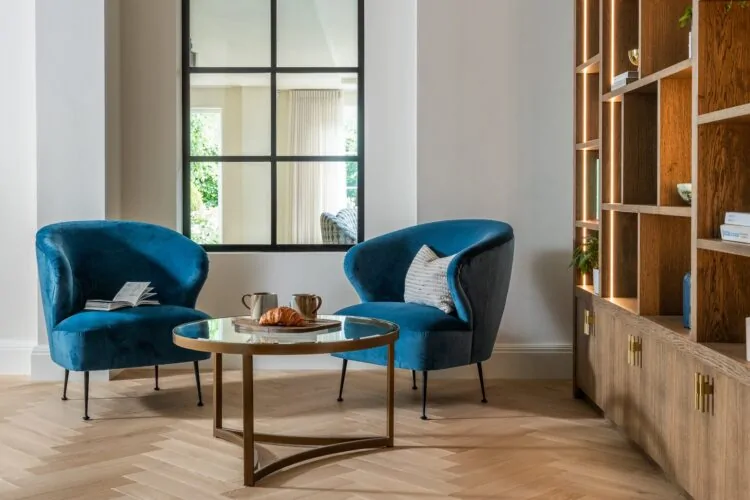 Two blue upholstered chairs sit facing a round glass table with a croissant and cups. A light wooden floor and a large window provide a bright, modern atmosphere.