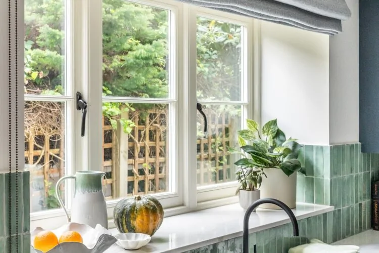A kitchen window features plants and fruits on a marble countertop beneath a light gray roller shade. Natural greenery is visible outside, creating a fresh, inviting atmosphere.