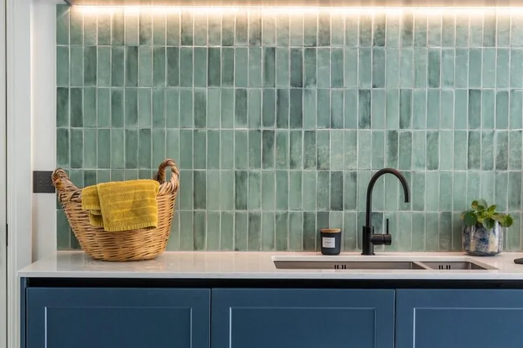 A wicker basket with a yellow towel sits on a countertop. Dual sinks and modern tap fixtures contrast against green tiled walls, illuminated by soft under-cabinet lighting in a sleek kitchen.