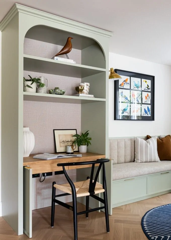 A green bookshelf with decorative items and plants surrounds a wooden desk and black chair, set against a light wall. A cozy, cushion-laden bench is nearby, enhancing comfort.