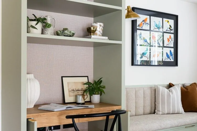 A green bookshelf with decorative items and plants surrounds a wooden desk and black chair, set against a light wall. A cozy, cushion-laden bench is nearby, enhancing comfort.
