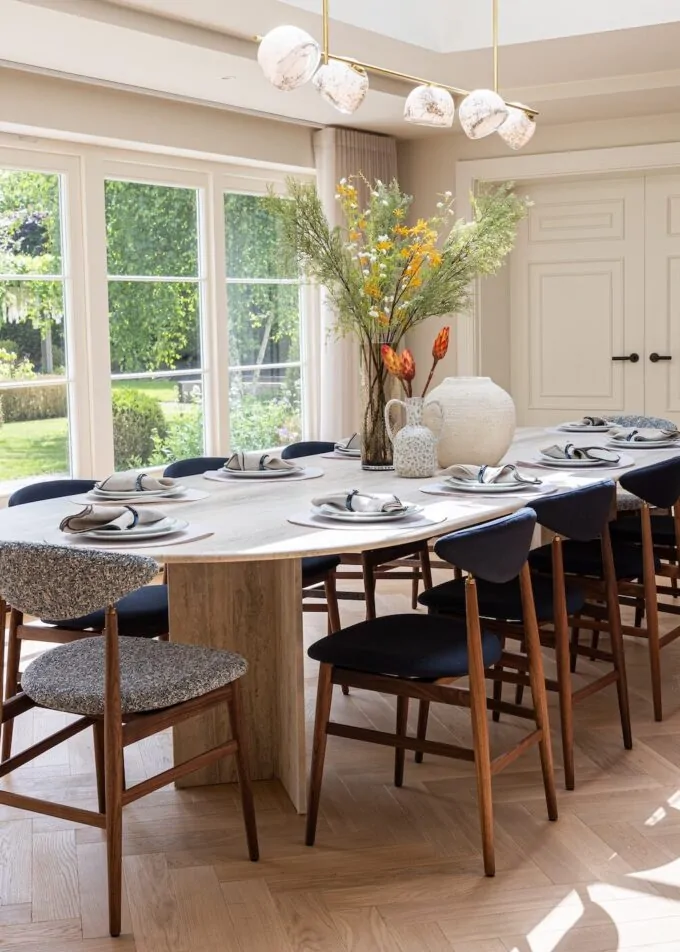 A round dining table is set with plates and cutlery, surrounded by high-backed chairs. A decorative vase with flowers is central, amidst a bright, sunlit room with large windows.