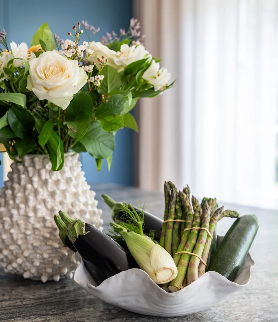 Art Deco Inspired Bespoke Kitchen made by our bespoke joiners, designed by Pfeiffer Design. Bespoke Joinery Kitchen with stone top island. Bunch of flowers in a spiky white ceramic vase and a selection of vegetables in a wavy ceramic white bowl