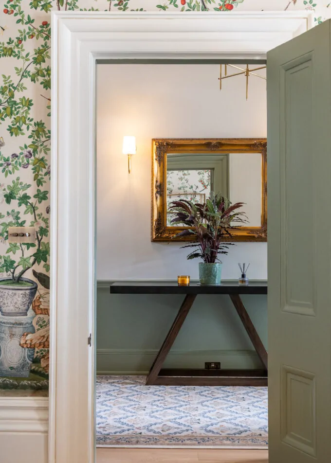 View through interior doorway into the corridor - looking at wooden console table and gold framed mirror with wall lights and plants