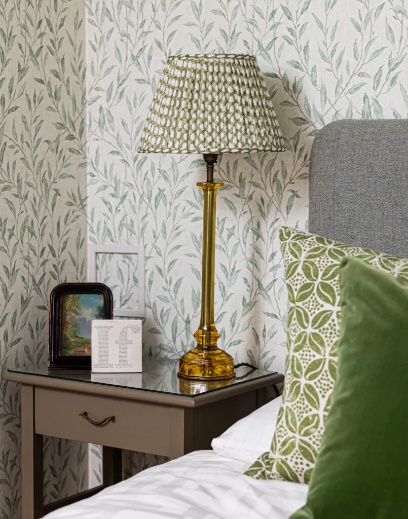 Green pleated lamp shade on bedside table blending with green floral wallpaper behind and green bedding