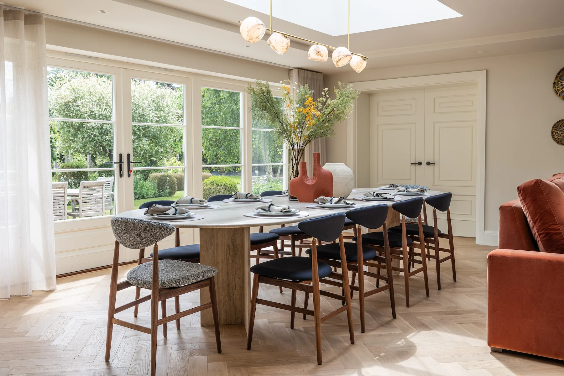 A round dining table is set with plates and cutlery, surrounded by high-backed chairs. A decorative vase with flowers is central, amidst a bright, sunlit room with large windows.